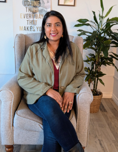 A smiling woman sitting in a cozy, modern chair in a well-lit office setting. She is wearing a green blazer over a maroon top, with jeans and a relaxed yet professional demeanor. A motivational quote is visible in the background, along with a vibrant indoor plant. The image highlights her approachable and professional presence, suitable for clinical supervision, corporate talks, speaking engagements, and consultations